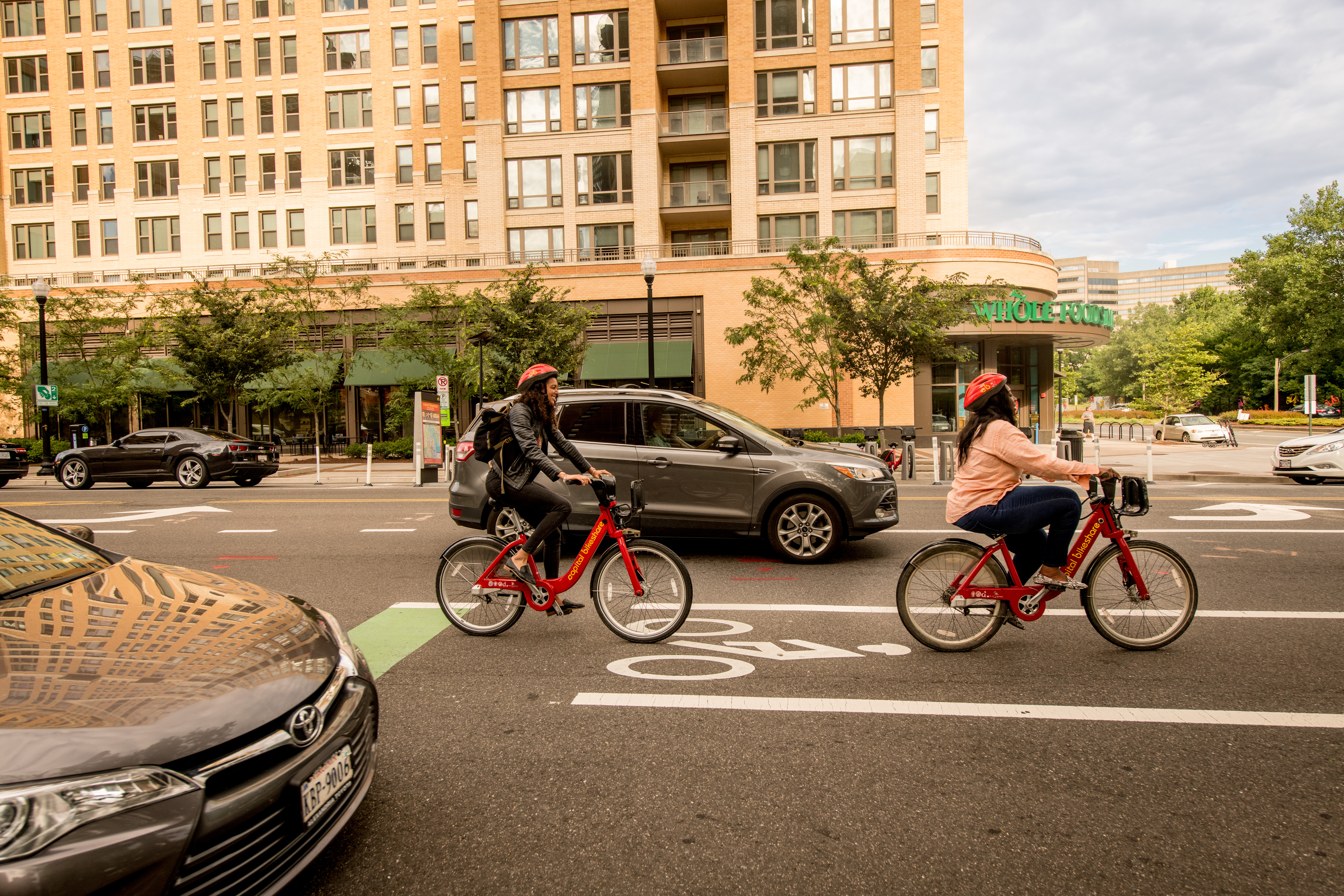 Two people riding on a red Capital Rideshare bikes passing in front of a residential building.